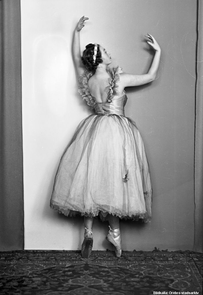 Kvinna i vit balettklänning och -skor står i en balettposition med ryggen mot kameran och ansiktet i profil.   