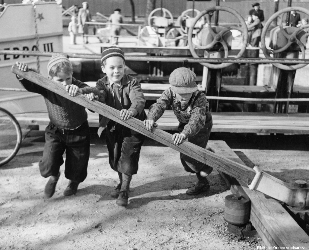 Tre barn skjuter en stor träpåle framför sig. I bakgrunden skymtar en båt i slussen.