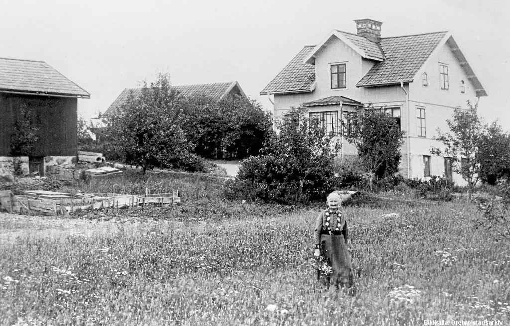En äldre kvinna står på en äng framför ett boningshus och två lador.