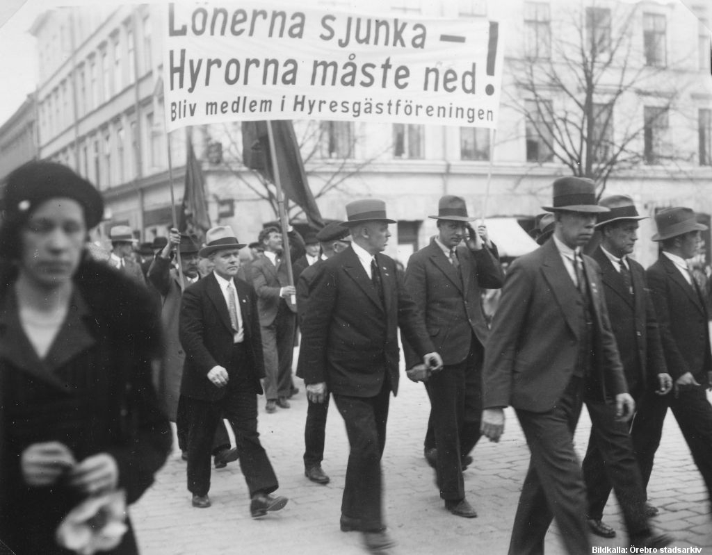 Hatt- och kostymklädda män går i grupp. På ett stort plakat står: "Lönerna sjunka - Hyrorna måste ned! Bliv medlem i Hyresgästföreningen"