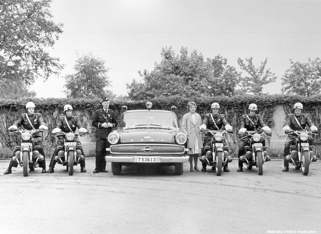 Två personer, en uniformsklädd och en civilklädd, står vid en polisbil. Två poliser på motorcyklar till vänster om bilen, tre poliser på motorcyklar till höger.