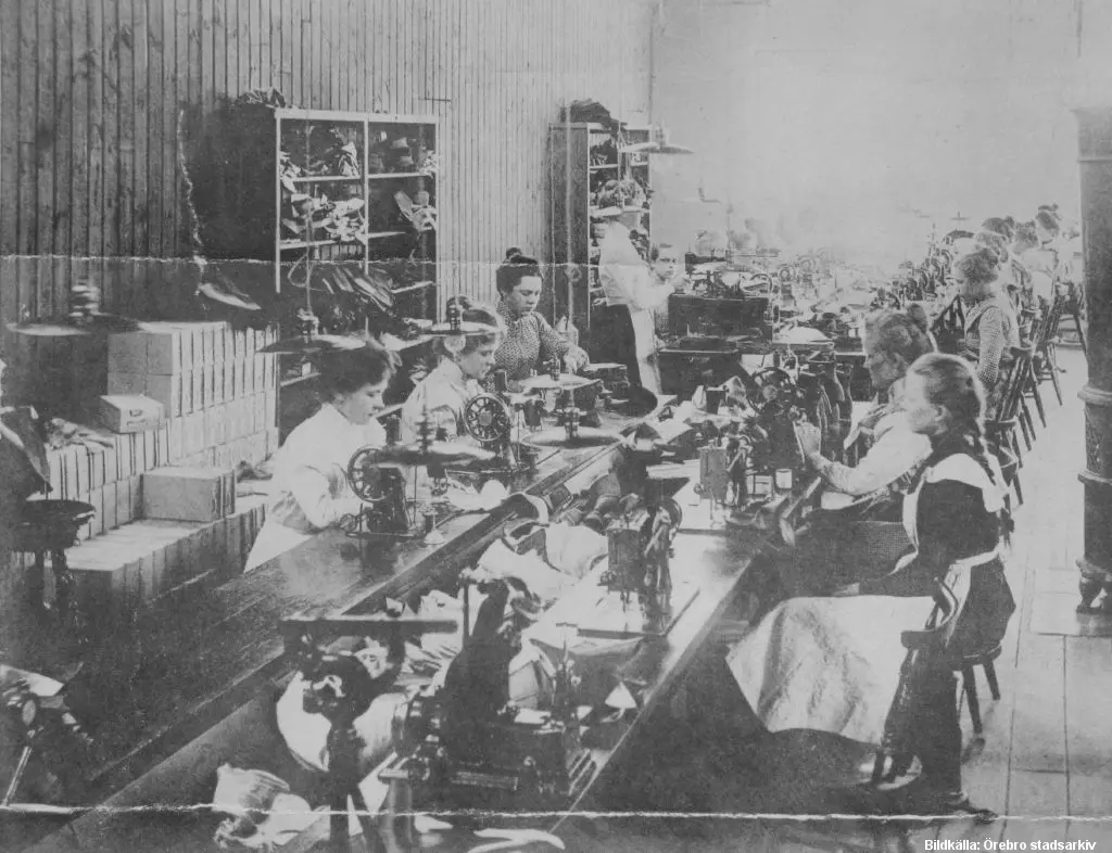 Långt bord med symaskiner där arbetare syr skor.