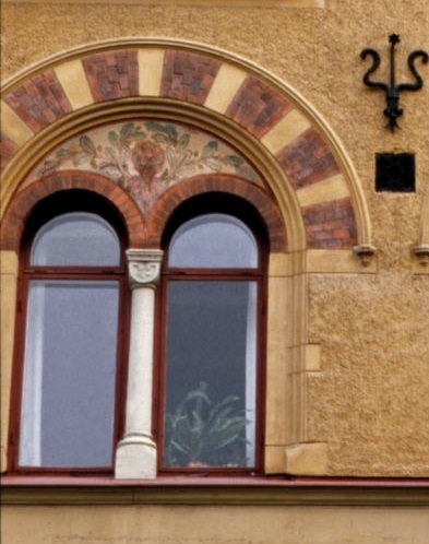 Ett dekorativt murat rött tegel under en halvmåneformad list möter den gula putsen. Den vita pelaren delar av de två rödmålade bågformade fönstren som kröns av en prydnadsgavel, dekorerad med ekblad och små ekollon.