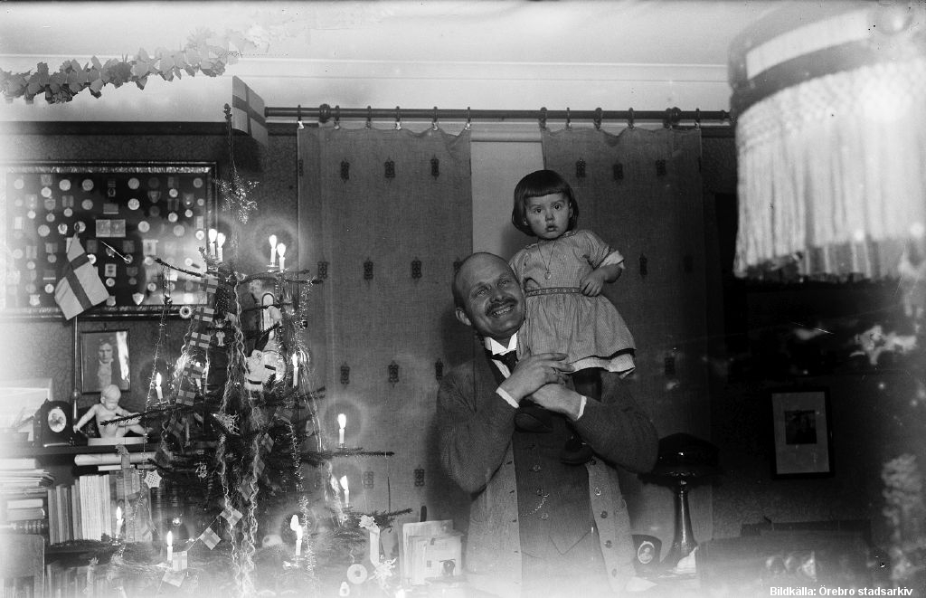 En man håller upp ett barn på sin axel. Till vänster om dem står en julgran.
