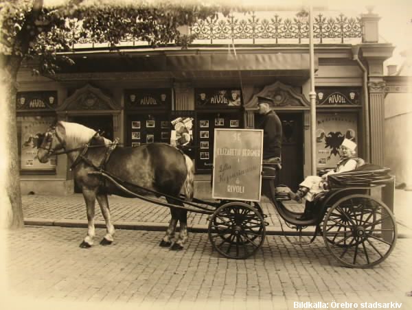 Framför entrén till en biograf står en vagn dragen av en häst. I vagnen sitter en kusk och en passagerare. Vagnen har en reklamskylt.