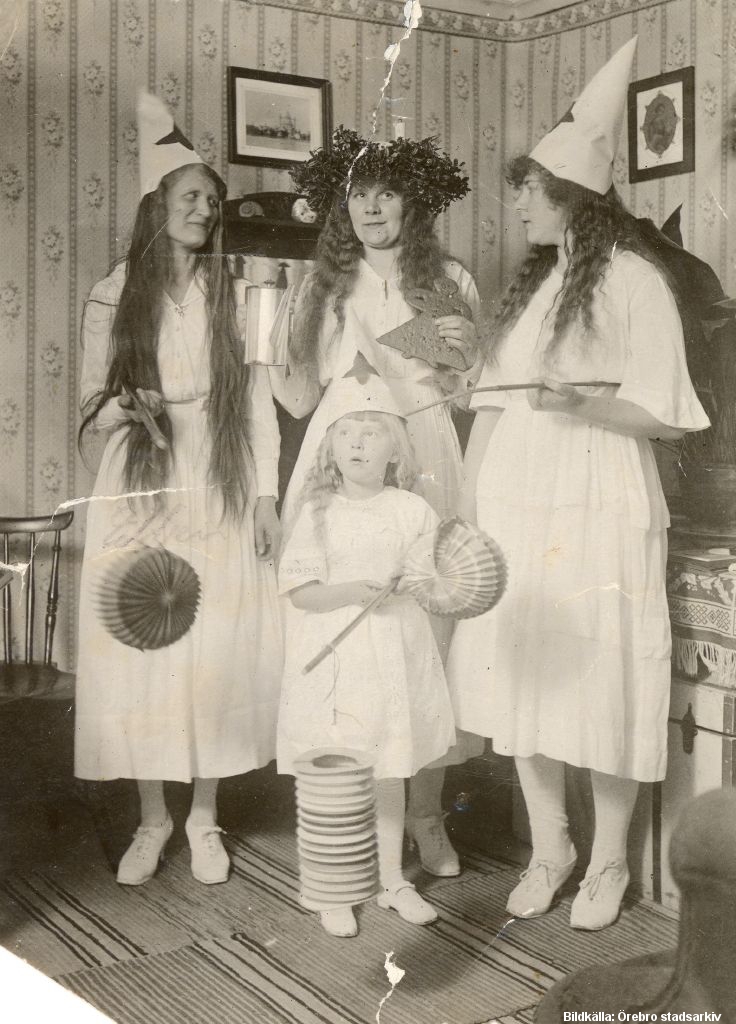 Tre kvinnor med långt hår i vita klänningar. Kvinnan i mitten har en krans i håret, de andra har vita strutar med stjärnor på. Framför dem står ett barn med vitt klänning och strut på huvudet.