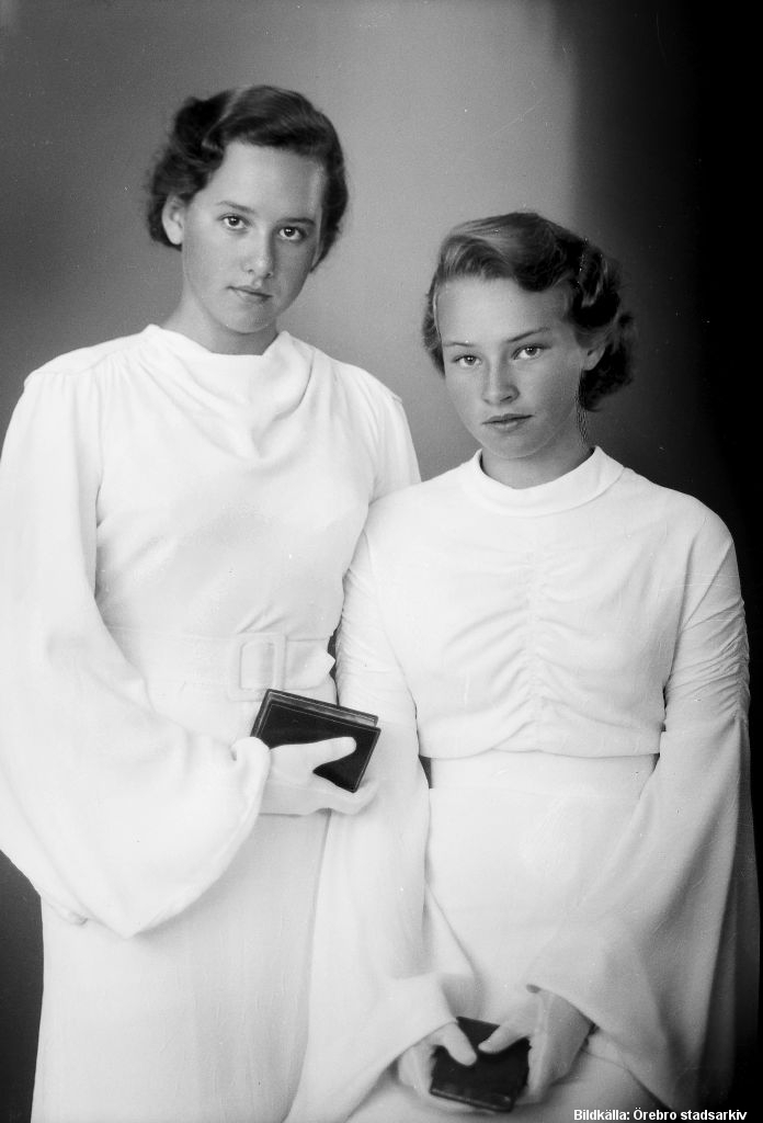Två flickor med vita klänningar och handskar. De håller i varsin svart bok.