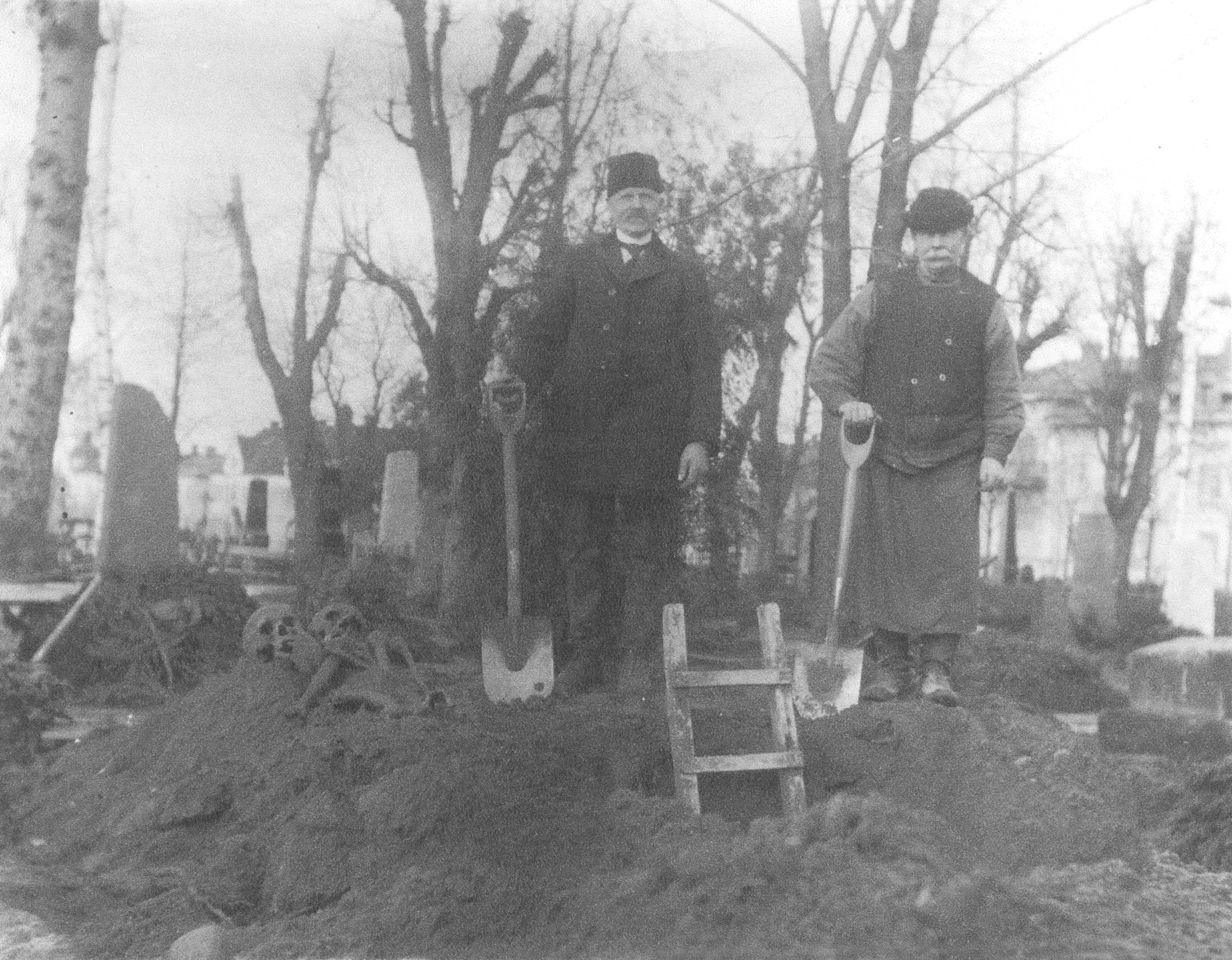 Två män står med spadar i jordhög. Gravstenar och träd skymtar i bakgrunden. 