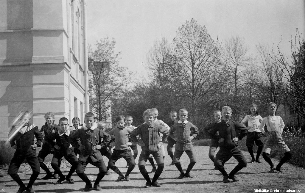 En grupp barn står utomhus, alla gör samma rörelser med armarna i sidorna och böjda ben.