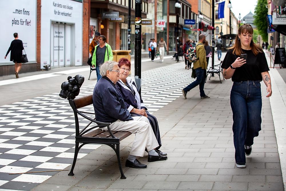 En man och en kvinna sitter på en parkbänk i stadsmiljö. En skulptur är placerad på ryggstödet. En kvinna går förbi framför dom. Skulpturen är cirka 30-40 cm hög och är gjord av hårt ihoplindad svart textil.