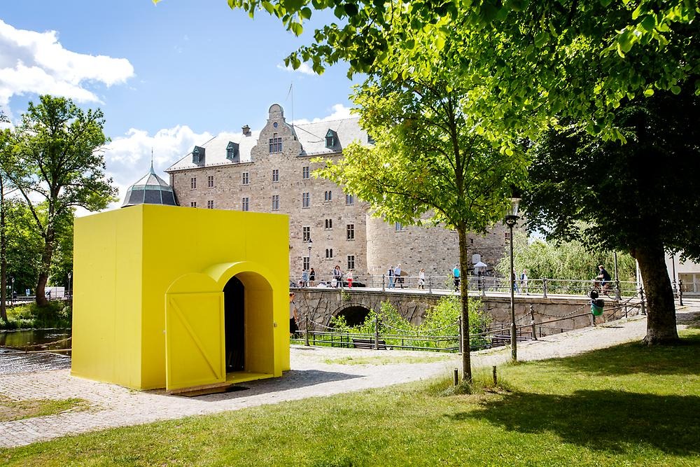 En gulmålal byggnad med välvd dörr är placerad på en avsats som vätter ut över Svartån, i bakgrunden syns Örebro slott, i förgrunden syns gräs och träd 