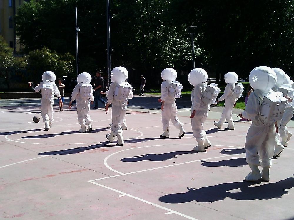Åtta människor som är utklädda till astronauter går över en basketplan, de har ryggarna mot oss som tittar.
