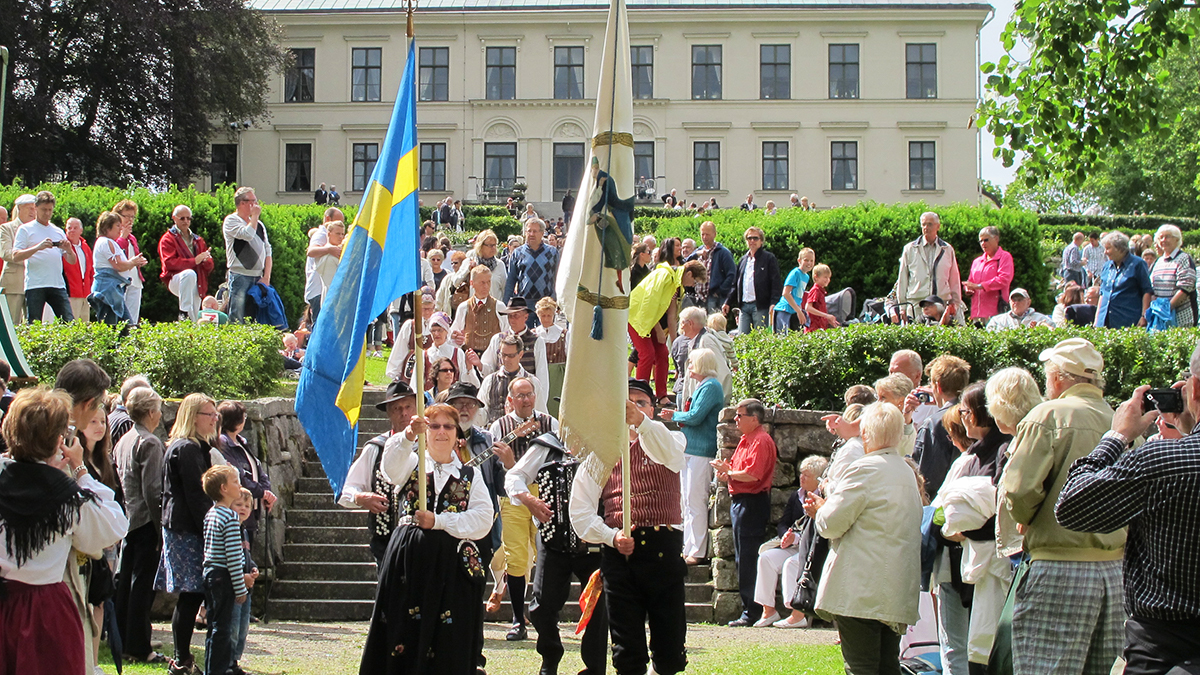 Två personer klädda i folkdräkt bär på fana och svenska flaggan. Besökare i bakgrunden