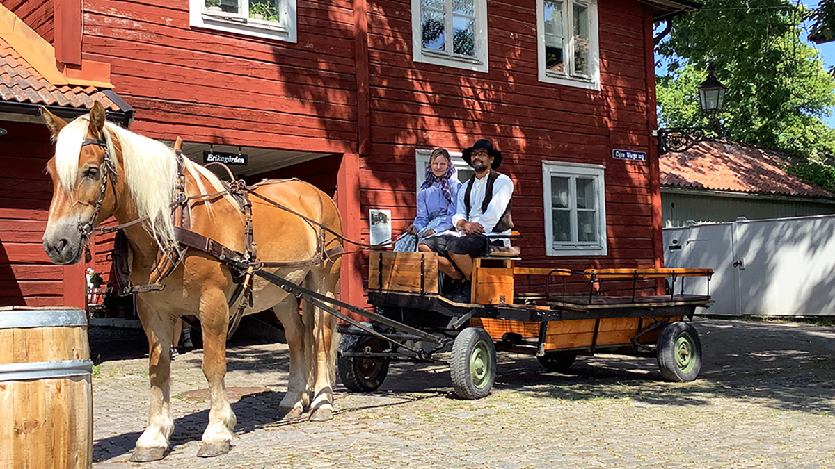 Häst och vagn. På vagnen sitter två personer i gammaldags kläder.