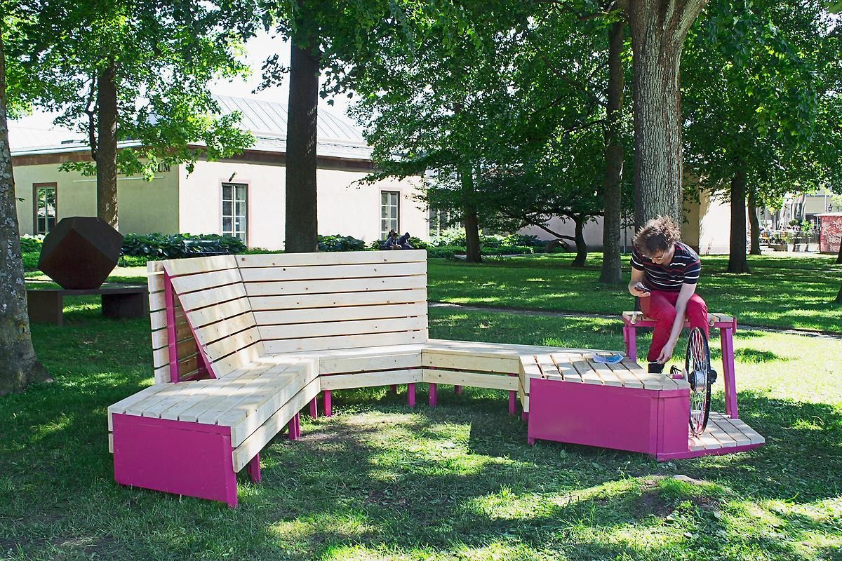 På en gräsplätt är en rosa och trädfärgad bänk placerad. Men detta är ingen vanlig bänk för på hörnet av den är bänken ihopkopplad med ett cykelhjul och två trampor så man kan skapa rörelseenergi vilket en kille sitter och gör på bilden.