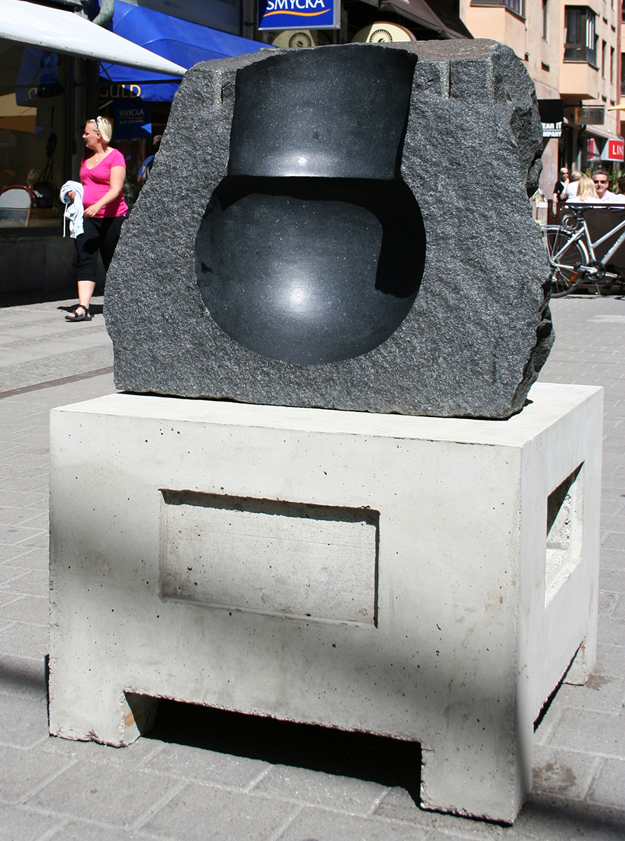 På ett betong bord utomhus står en skulptur som avgjutit en kettlebell liknande form. 