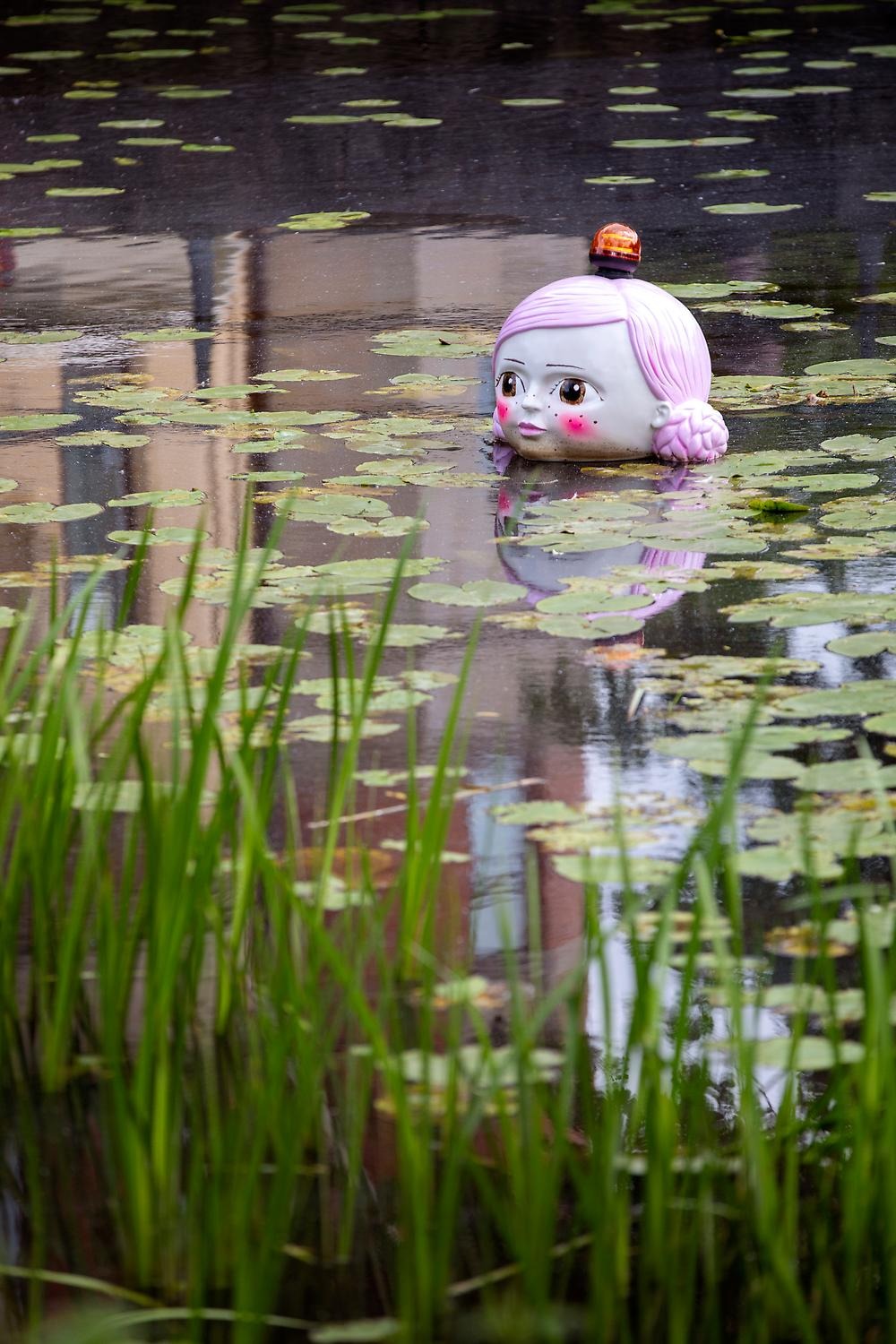 I vattnet bland näckrosor flyter ett stort flickansikte. Hon har rosa flätor och en lampa på huvuvdet.