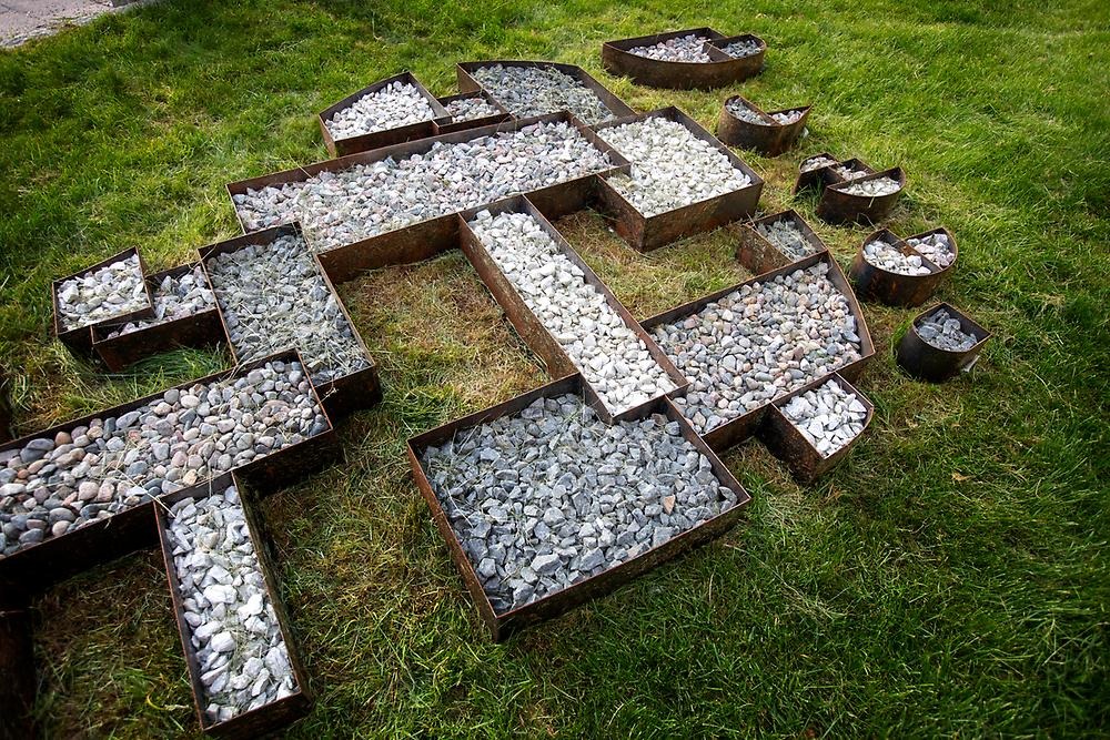 The picture shows outdoors, a lawn, on the lawn are containers of metal placed, in the containers there are stones. The containers together form a pattern that shapes the upper part of a foot, with five toes.