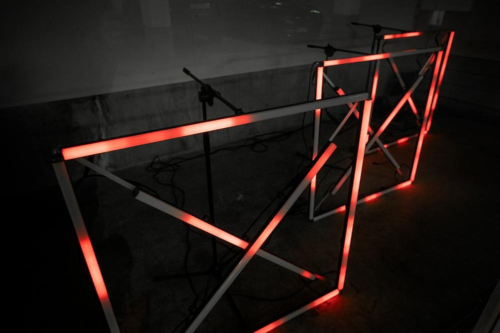 Innanför glasrutorna i ett utställningsrum i ett garage syns tre stycken fyrkantiga moduler med ett kryss i varje modul. Det är led-lampor som ger hela rummet ett rött sken. Bilden visar en närbild på de tre modulerna.