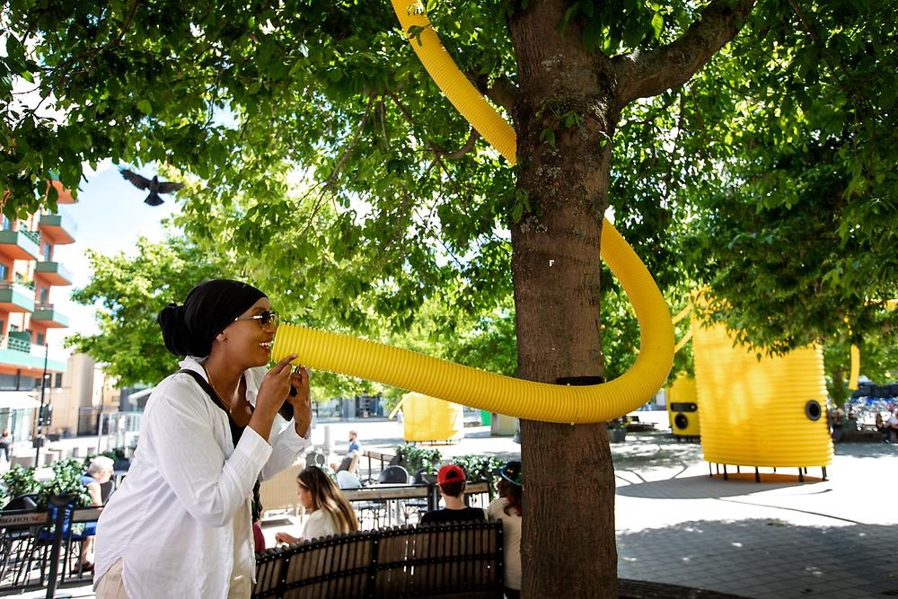 En kvinna med solglasögon, slöja och vit skjorta ser glad ut när hon pratar in i ett gult dräneringsrör i en torgmiljö. Röret kommer uppifrån i bild och är lindat runt ett träd. I bakgrunden syns torget med konstverket i bakgrunden. 