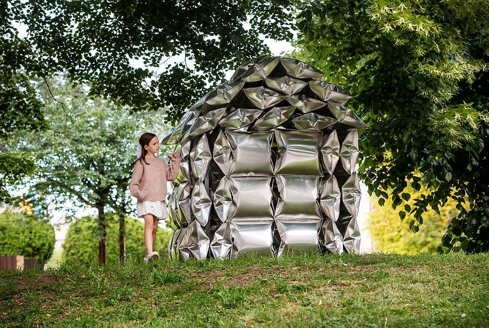 I en lummig park står ett hus som är gjort av högblanka aluminumkuddar. Omgivningen runtomkring speglas i kuddarna. En flicka står bredvid huset/skulpturen.