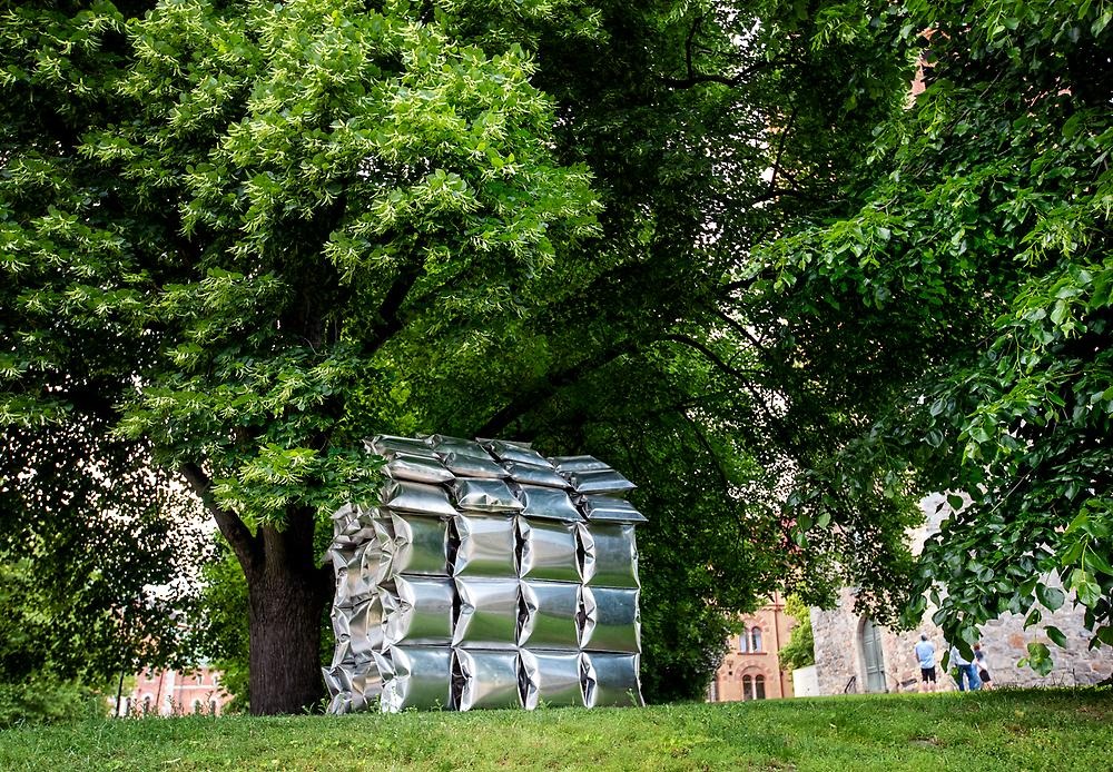 I en lummig park står ett hus som är gjort av högblanka aluminumkuddar. Omgivningen runtomkring speglas i kuddarna.