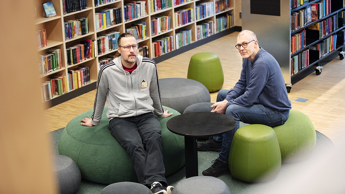 Två män sitter i ett bibliotek med blicken i kameran