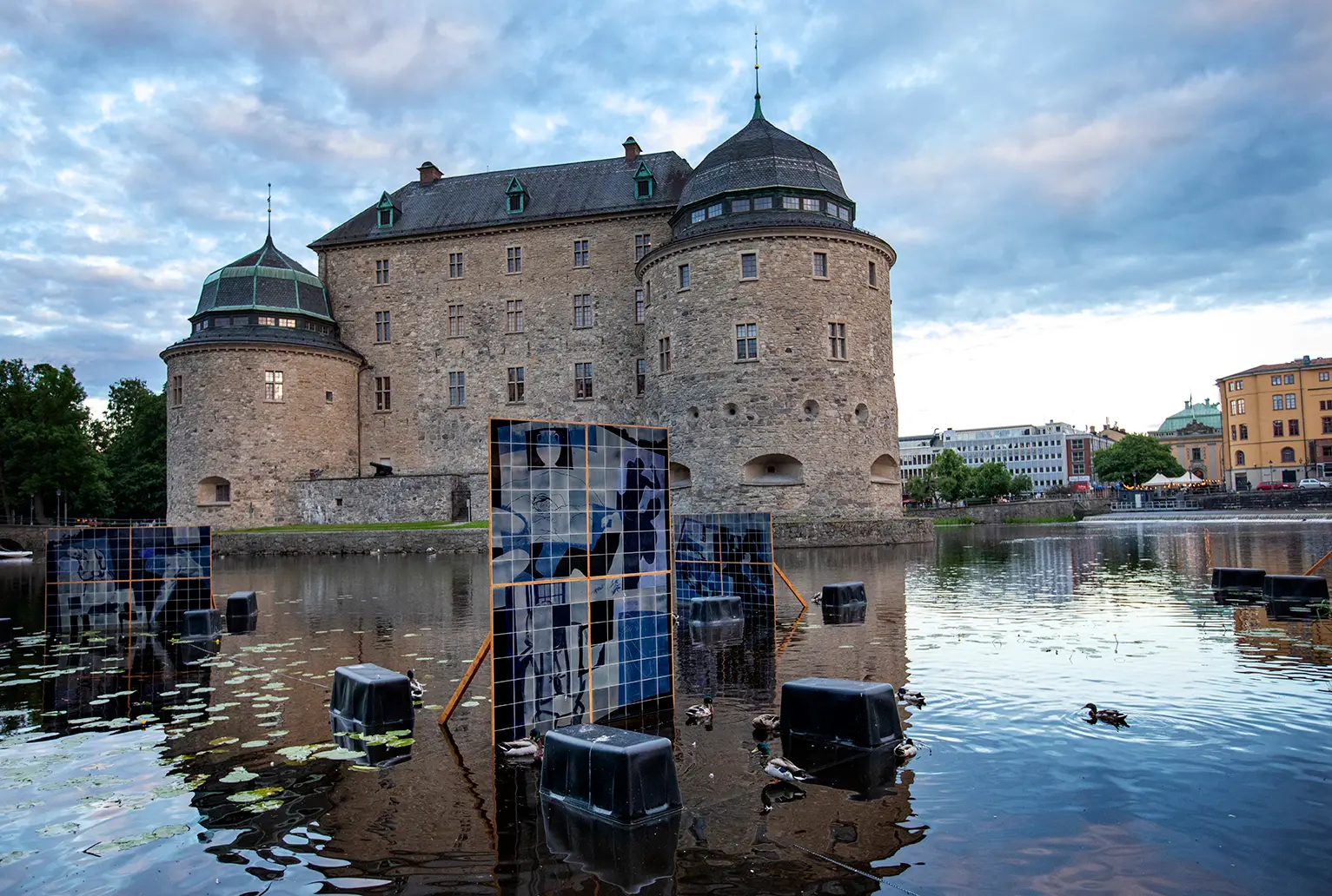 Översiktsbild på tavlor gjorda av keramikplattor som står placerade i vattnet runt Örebro slott. Keramikplattorna går i olika nyanser av blå och på dem syns manskroppar.