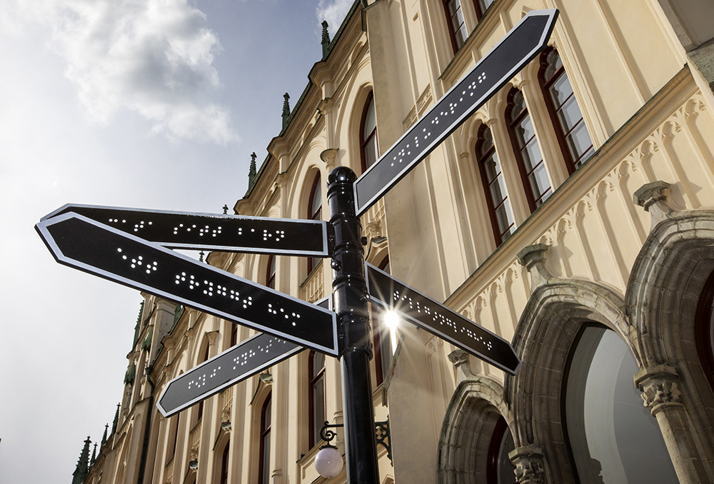 In the foreground of the City Hall in Örebro, a sign is used that is usually used as a directions sign. Instead of the Latin alphabet, Braille can be seen on the signs. The sign is black with an arrow formation at the front. A white border can be seen around the outer edges of the sign. The image is a close-up of the signs pointing in different directions.